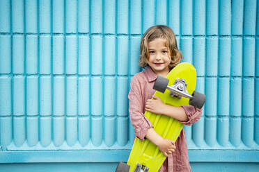 Cute girl holding skateboard while standing in front of blue wall - RCPF01272