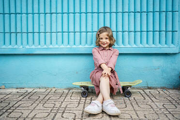 Smiling girl sitting with legs crossed at knee on skateboard in front of blue wall - RCPF01273