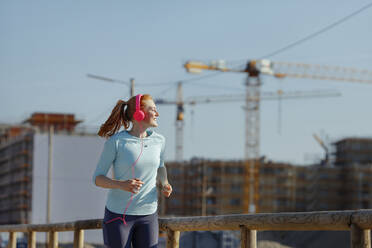 Lächelnde Frau beim Joggen in der Nähe einer Baustelle an einem sonnigen Tag - AANF00069