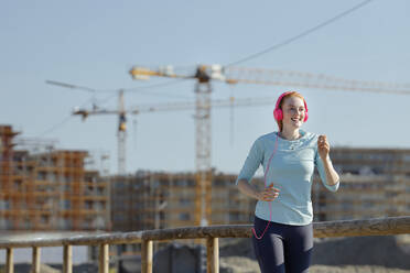 Junge Frau mit Kopfhörern beim Joggen auf einer Baustelle - AANF00070