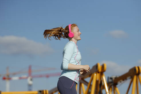 Lächelnde Frau mit Kopfhörern beim Joggen an einem sonnigen Tag - AANF00079