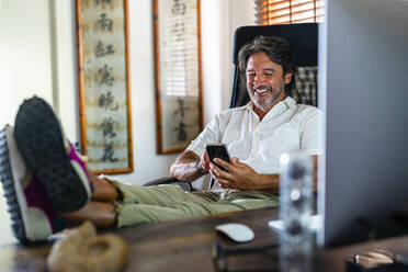 Smiling male professional using mobile phone while relaxing on chair at home office - DLTSF02189