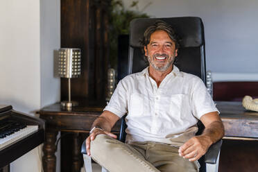 Smiling mature man sitting on chair at home - DLTSF02190