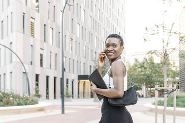 Happy female business professional talking on mobile phone while walking in city - JCZF00838