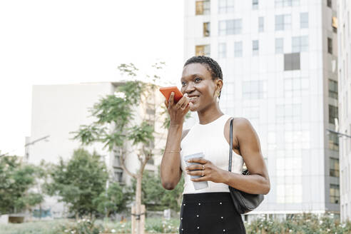 Female freelancer holding coffee cup while sending voicemail through smart phone in city - JCZF00842