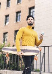 Mann mit Skateboard und Mobiltelefon geht an einem Gebäude vorbei - JCCMF03864