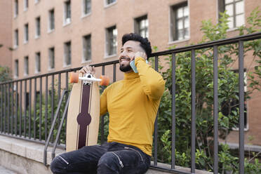 Lächelnder gutaussehender Mann mit Skateboard, der mit seinem Handy telefoniert - JCCMF03869