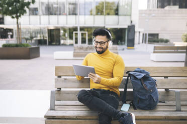 Happy man with headphone using digital tablet on bench - JCCMF03884