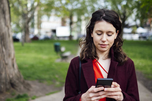 Junge Frau, die im Park eine Textnachricht über ihr Mobiltelefon sendet - AMWF00002