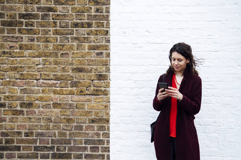 Frau surft mit ihrem Smartphone im Internet, während sie vor einer Backsteinmauer steht - AMWF00004