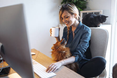 Smiling female influencer looking at dog while having coffee at home - JSRF01627