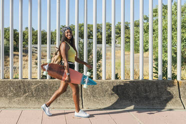 Smiling woman with backpack and skateboard walking on footpath - JRVF01799