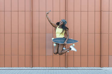 Smiling woman with skateboard jumping while taking selfie through smart phone in front of wall - JRVF01804