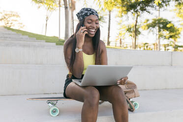Happy woman talking on mobile phone while sitting with laptop on skateboard - JRVF01817