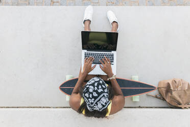 Frau mit Kopftuch benutzt Laptop, während sie auf einem Skateboard sitzt - JRVF01818