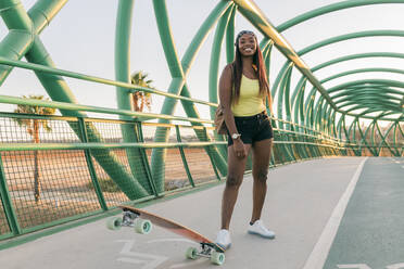 Lächelnde Frau steht mit Skateboard auf einer Brücke - JRVF01825