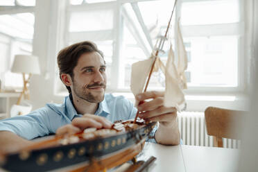 Männlicher Geschäftsmann betrachtet den Mast eines Spielzeugboots im Büro - KNSF08960