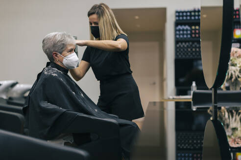 Friseurin mit Gesichtsmaske beim Schneiden der Haare eines Kunden im Salon - OCAF00766