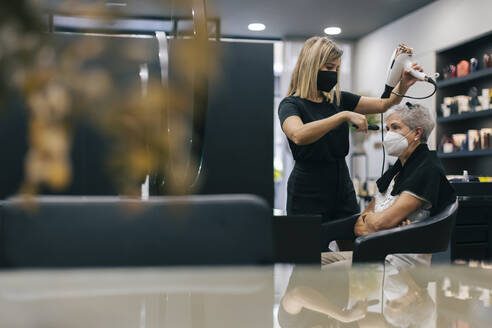 Friseurin mit Gesichtsmaske föhnt die Haare eines Kunden im Salon - OCAF00767