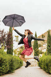 Glückliche erwachsene Frau, die bei Regen im Park mit einem Regenschirm springt - MGRF00496