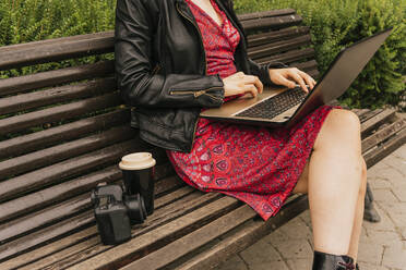 Junge Fotografin mit Laptop, während sie mit Kamera und Einwegbecher auf einer Bank im Park sitzt - MGRF00497