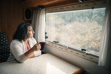 Frau mit Kaffeetasse schaut durch ein Wohnmobilfenster - GRCF00880