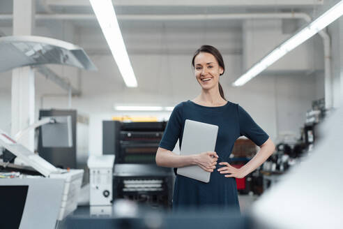 Happy businesswoman holding digital tablet while standing with hand on hip in industry - KNSF09026