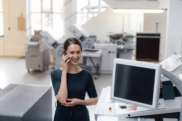 Lächelnde weibliche Fachkraft, die in einer Werkstatt mit einem Smartphone spricht - KNSF09045