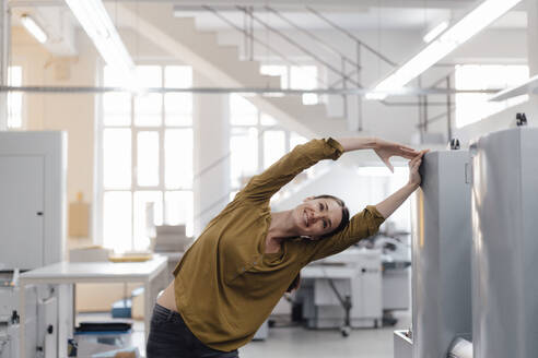 Smiling businesswoman stretching while exercising in industry - KNSF09047