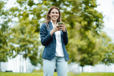 Glückliche junge Geschäftsfrau mit Smartphone in einem öffentlichen Park - GUSF06499