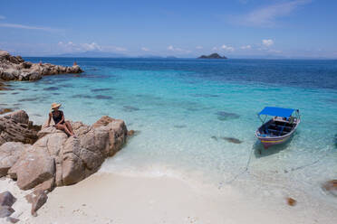 Weiblicher Reisender mit Hut sitzt auf Steinen am Sandstrand des klaren türkisfarbenen Meeres in Malaysia - ADSF30311