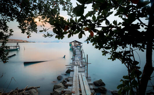 Blick durch die Äste der Bäume auf den Pier, der zu einer Hütte mitten im Meer führt, am Abend in Malaysia - ADSF30318