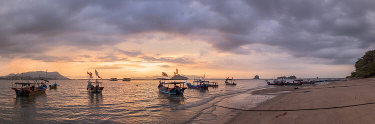 Weitwinkel von Fischerbooten mit wehenden Nationalflaggen auf nassen sandigen Küste von Meer unter düsteren bewölkten Himmel bei Sonnenuntergang in Malaysia gewaschen - ADSF30321