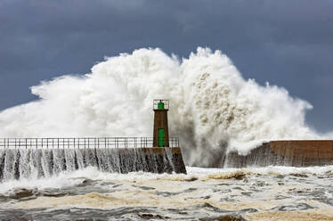 Huge foamy sea waves crashing against stony breakwater with old lighthouse tower against blue cloudy sky in Port of Viavelez in Asturias Spain - ADSF30325