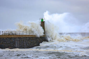 Huge foamy sea waves crashing against stony breakwater with old lighthouse tower against blue cloudy sky in Port of Viavelez in Asturias Spain - ADSF30326