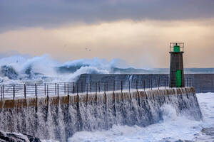 Riesige schäumende Meereswellen brechen gegen steinige Wellenbrecher mit altem Leuchtturm Turm gegen blauen bewölkten Himmel im Hafen von Viavelez in Asturien Spanien - ADSF30327