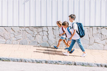 Side view of schoolboy with backpack speaking with female friends while strolling on tiled pavement against stone wall in sunlight - ADSF30335