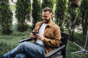 Lächelnder Geschäftsmann, der im Hinterhof eine Textnachricht über sein Handy verschickt - VPIF04904