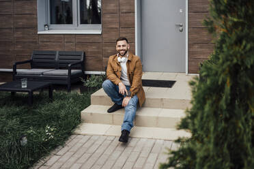 Lächelnder mittelgroßer erwachsener Mann sitzt auf den Stufen im Hinterhof - VPIF04908