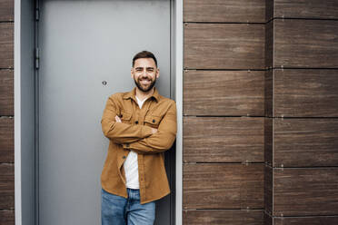 Smiling handsome man standing with arms crossed while leaning at doorway - VPIF04910