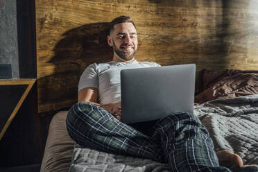 Smiling man using laptop on bed at home - VPIF04916