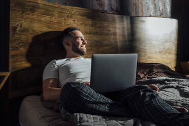 Smiling mid adult man with laptop on bed at home - VPIF04917