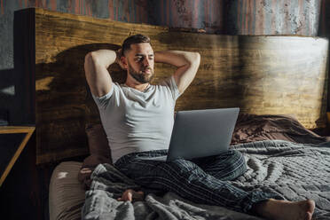 Handsome man with hands behind head looking at laptop while sitting in bedroom - VPIF04920