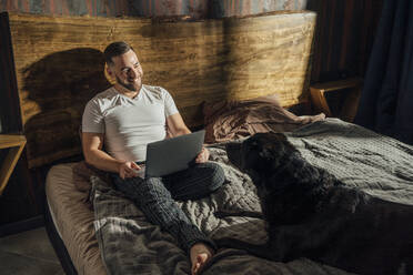 Lächelnder Mann mit Laptop, der den Hund auf dem Bett zu Hause betrachtet - VPIF04921