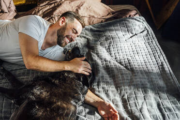 Happy man stroking dog on bed at home - VPIF04925