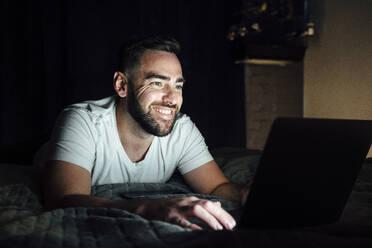 Glücklicher Mann mit Laptop auf dem Bett in einem dunklen Raum - VPIF04933