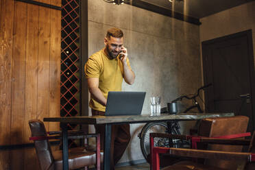 Smiling male professional using laptop while talking on smart phone at home office - VPIF04955