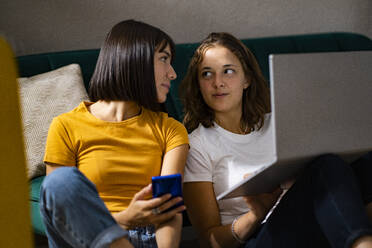 Lesbisches Paar mit Mobiltelefon und Laptop, die sich gegenseitig anschauen - GIOF13512