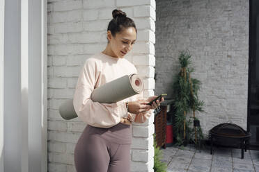 Mid adult woman with yoga mat using mobile phone on terrace - VPIF04982