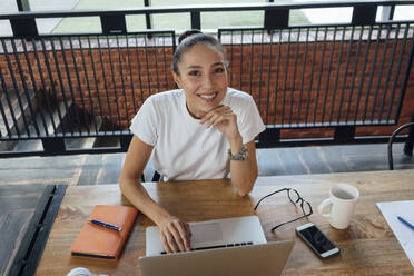 Smiling businesswoman with hand on chin sitting at desk in office - VPIF04995